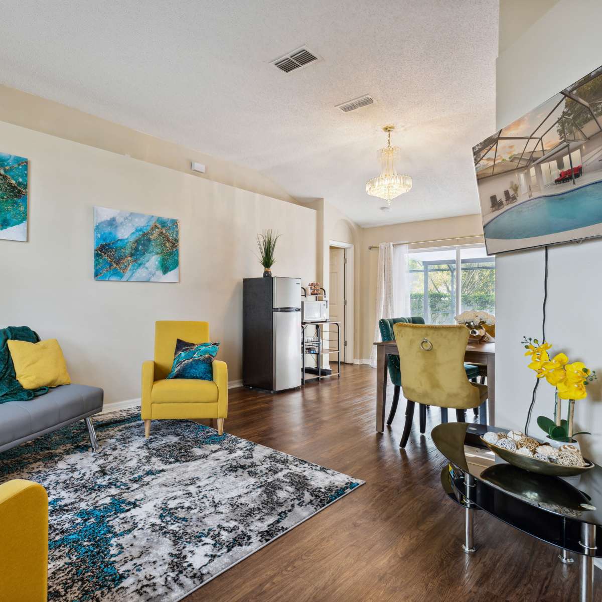 A modern living room features a gray sofa, yellow chairs, abstract artwork, and a dining area with a view of the outdoors.