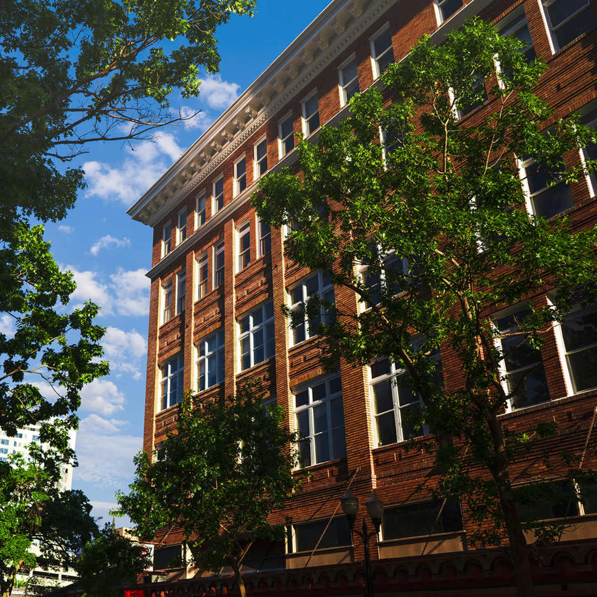 A red brick building with large windows is surrounded by green trees against a blue sky with scattered clouds.