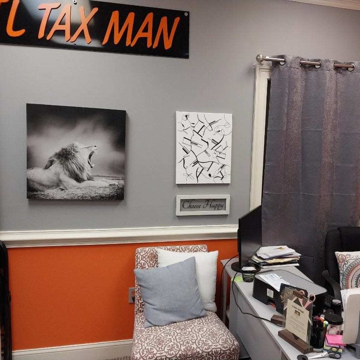 An office space with orange and gray walls featuring a lion photograph and abstract art, a cluttered desk with an open laptop, and a chair with decorative cushions.