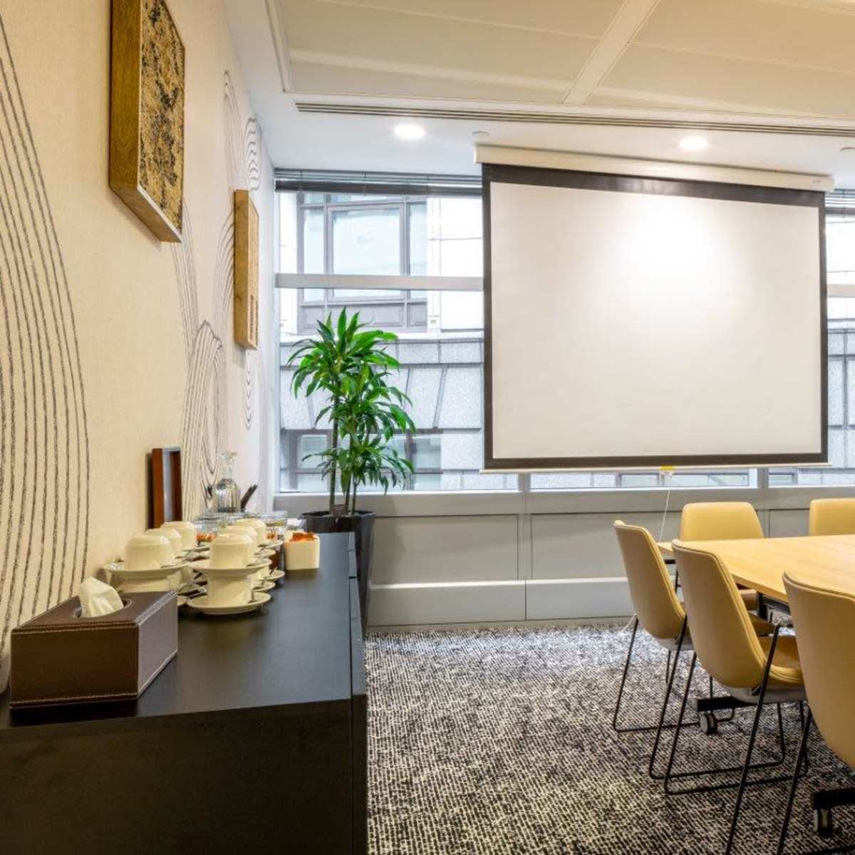 A conference room features a large wooden table surrounded by beige chairs, with a projector screen on one wall and a plant in the corner.