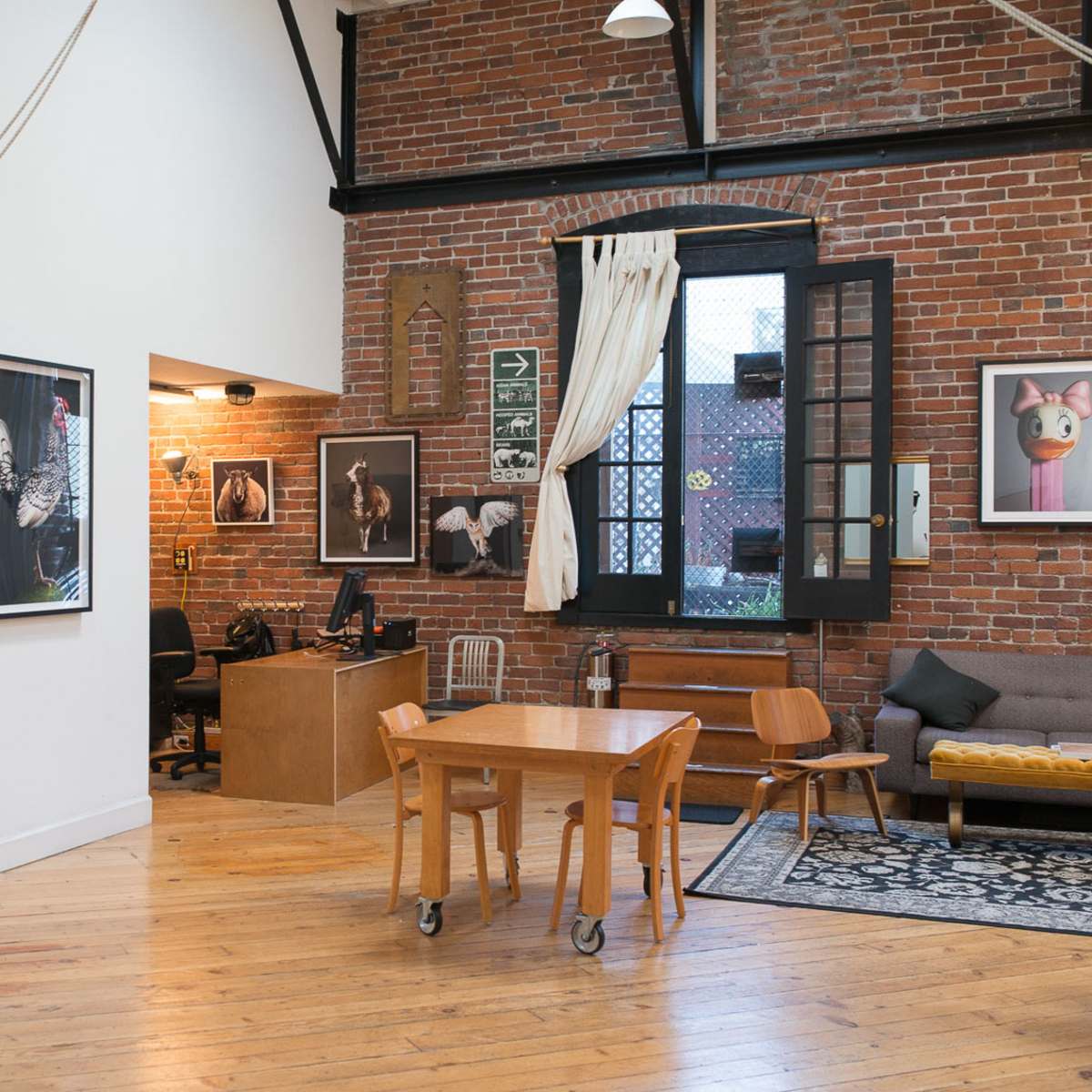 An open loft space with exposed brick walls, a wooden floor, a table and chairs, and a workspace featuring artwork on the walls.