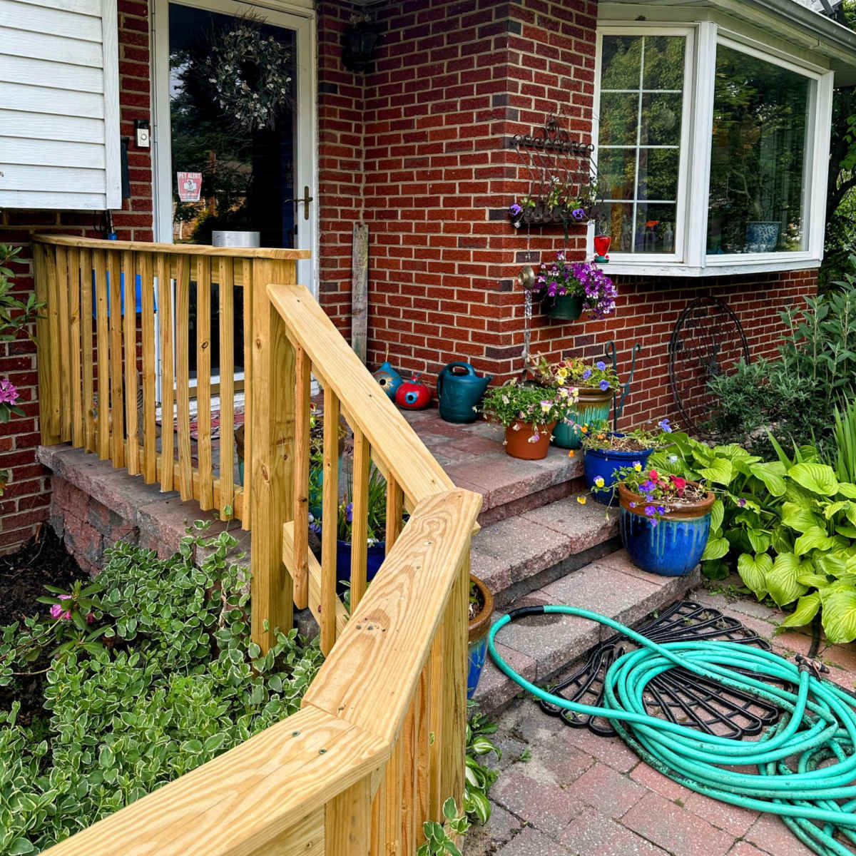 A brick house entrance with a wooden ramp, surrounded by flower pots and greenery.