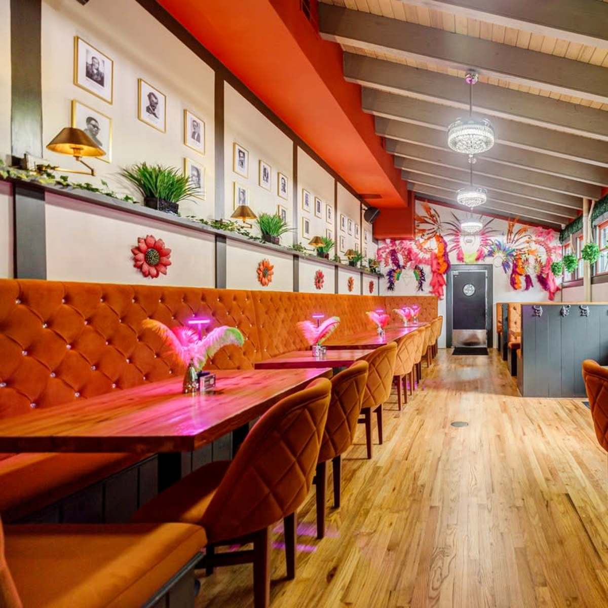 The interior of a restaurant featuring wooden tables, orange upholstered chairs, and decorative plants along the walls, with colorful lighting and framed photographs.