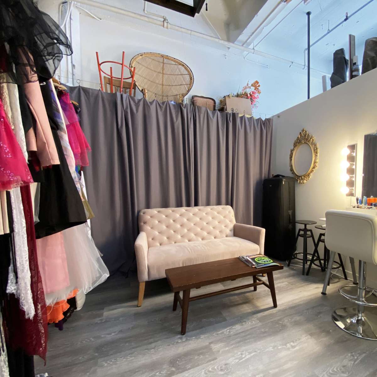 A dressing room with a pink sofa, a wooden coffee table, a vanity with bright lights, and clothing hanging on the wall behind a gray curtain.