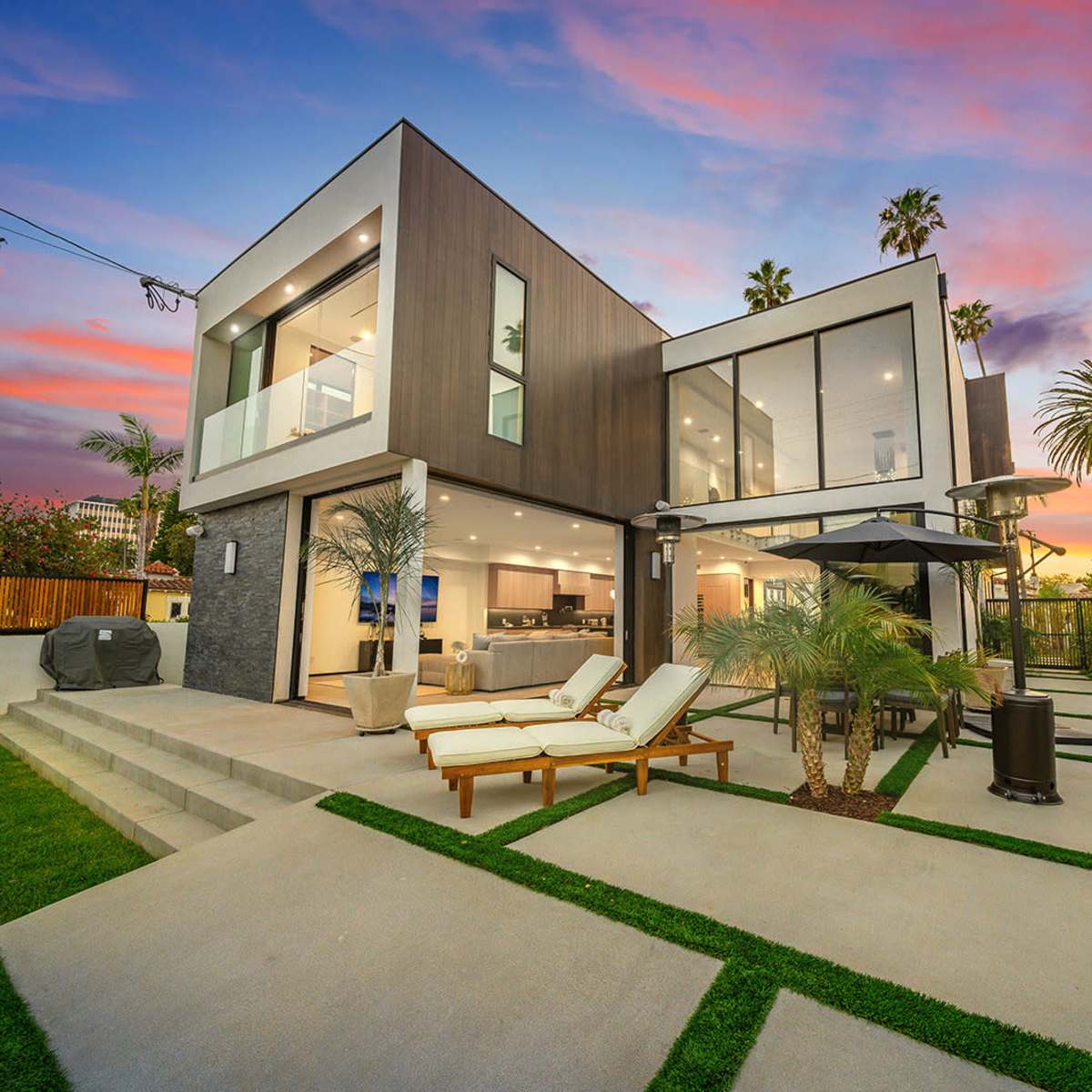 A modern two-story house with large glass windows and a landscaped backyard is set against a colorful sunset sky.