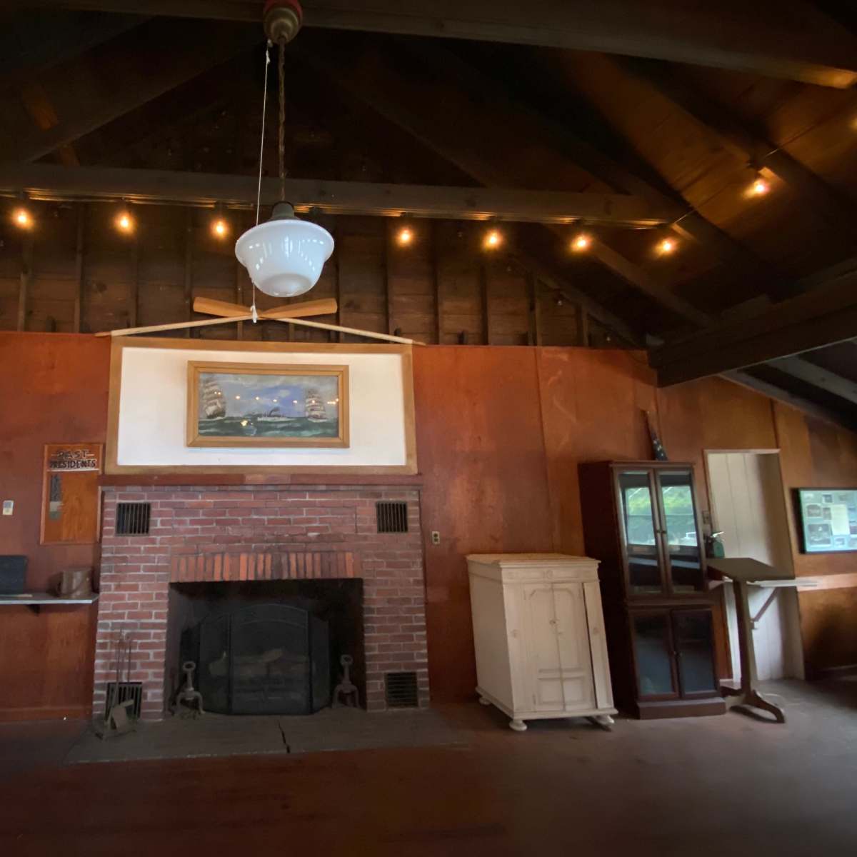 The interior of a rustic room features a brick fireplace, wooden beams, and a painting above the mantle.