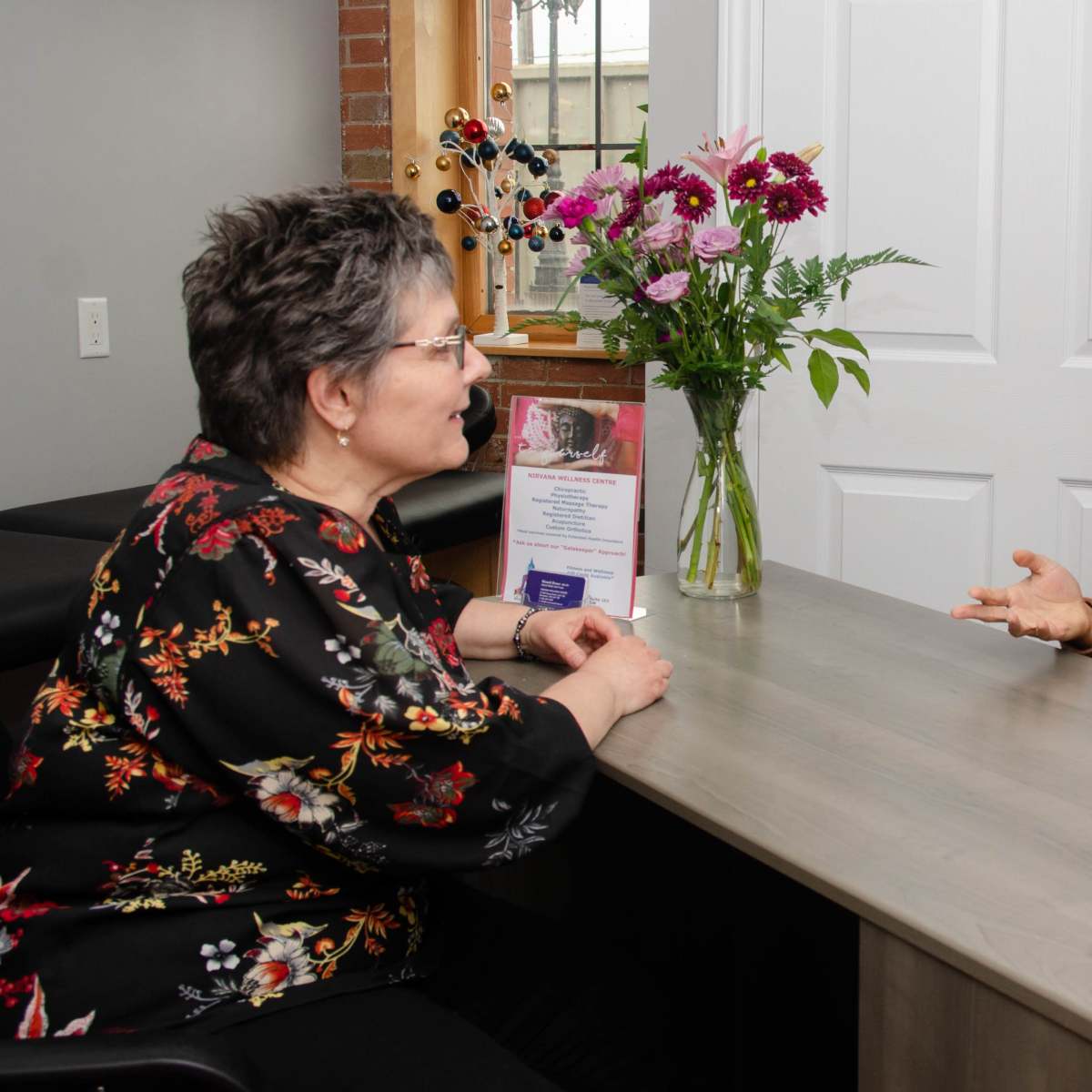 A woman with curly hair speaks to another woman seated at a desk with flowers and promotional materials in the background.