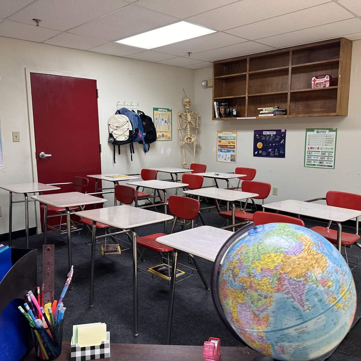 A classroom with several empty red chairs and desks, a globe, a skeleton model on the wall, and educational posters.