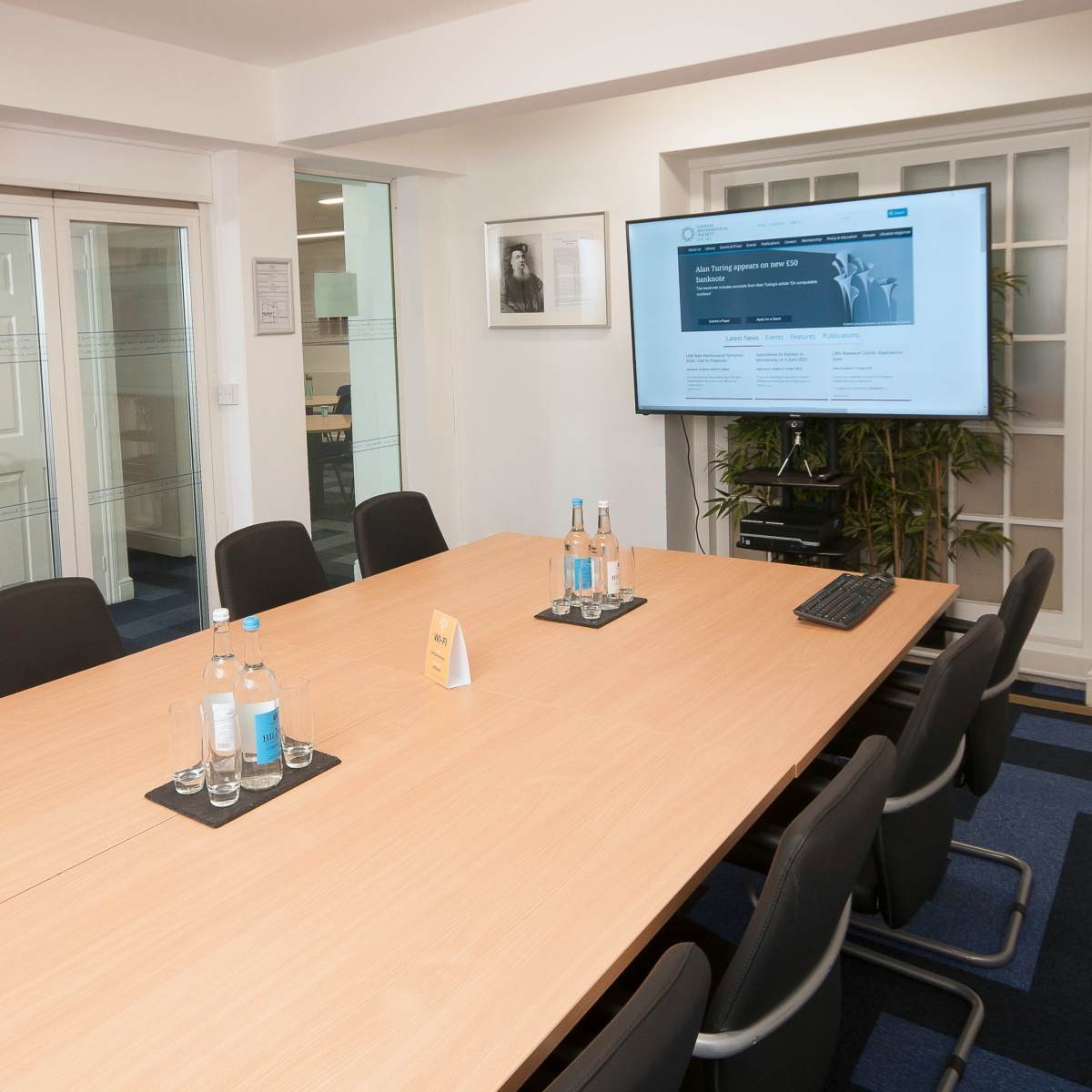 A modern conference room features a long wooden table, black chairs, and a television screen displaying a webpage.