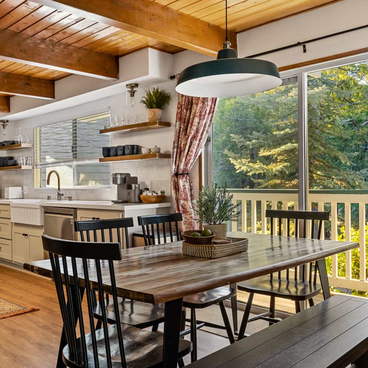 A modern kitchen features wooden beams, a large dining table, and a view of greenery through a sliding glass door.