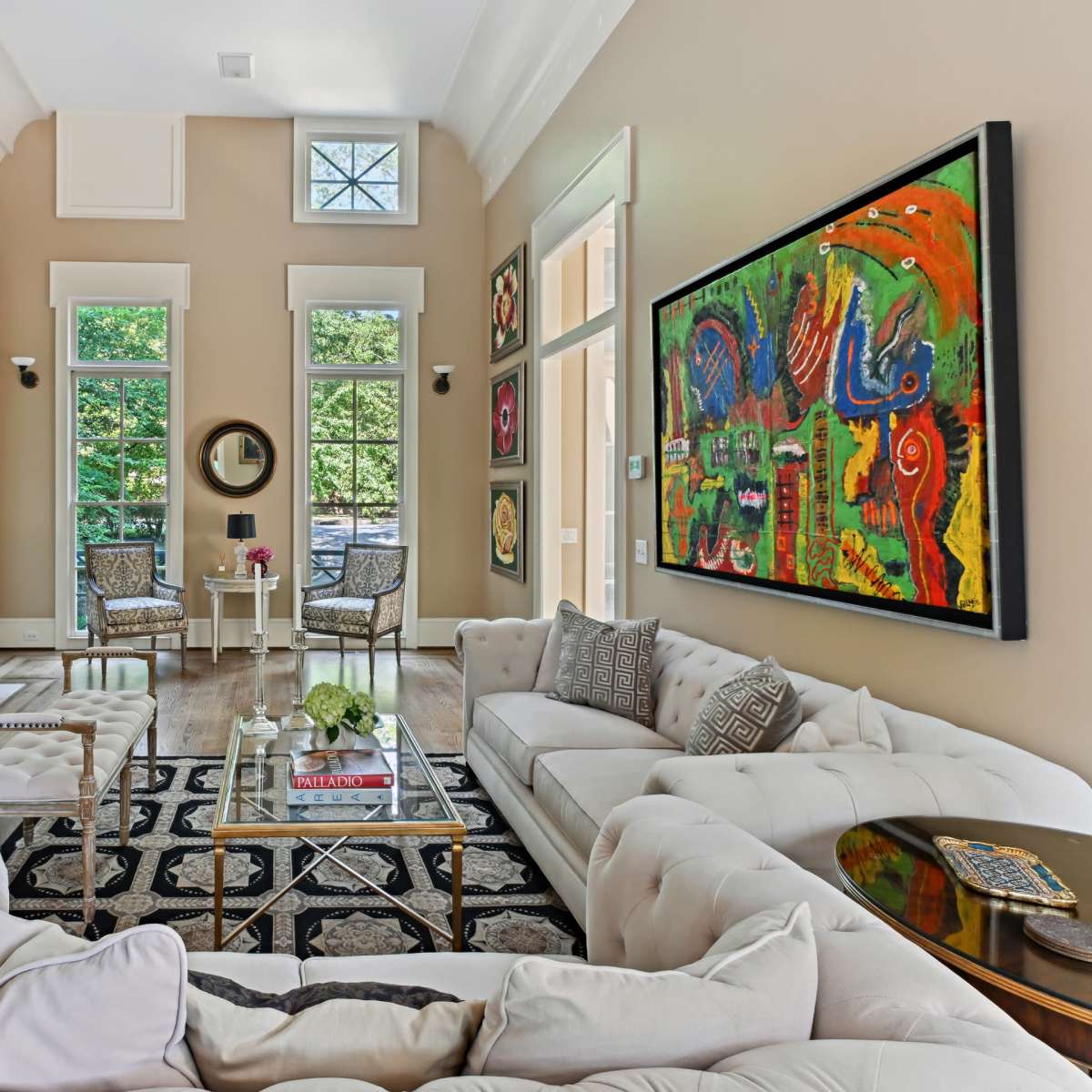A well-lit living room featuring a large artwork on the wall, a white sofa, a coffee table in the center, and large windows overlooking greenery.