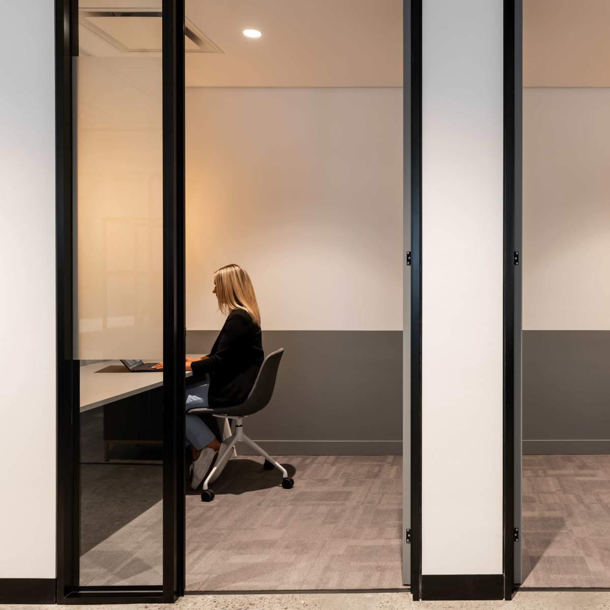 Two glass-walled office cubicles contain individuals sitting at desks, with one person visible in each space.