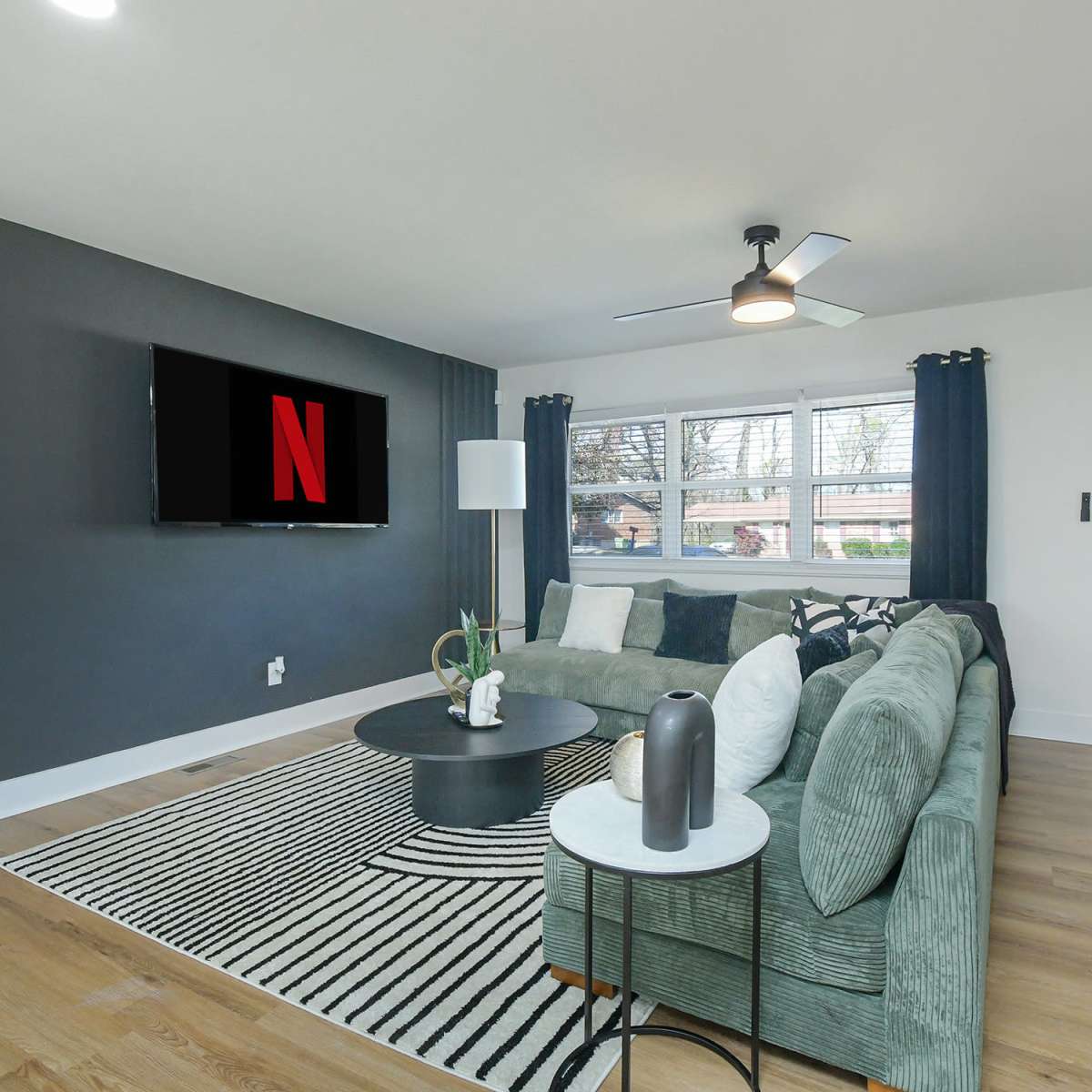The living room features a green sectional sofa, a round coffee table, a TV mounted on the wall displaying the Netflix logo, and large windows with black curtains.