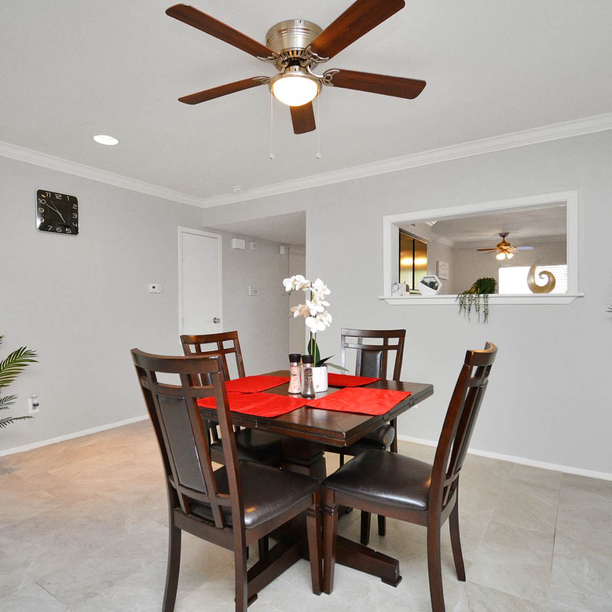 A dining area featuring a wooden table with four chairs, a ceiling fan, and decorative plants, set against light gray walls.