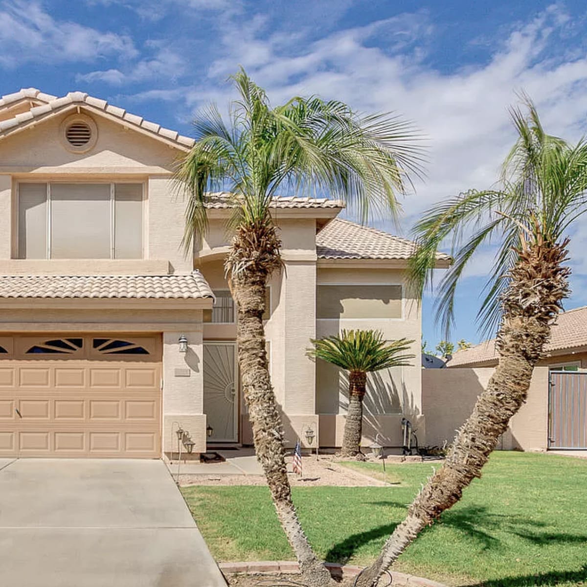 A two-story beige house with a tiled roof, palm trees in the front yard, and a two-car garage is situated under a blue sky.