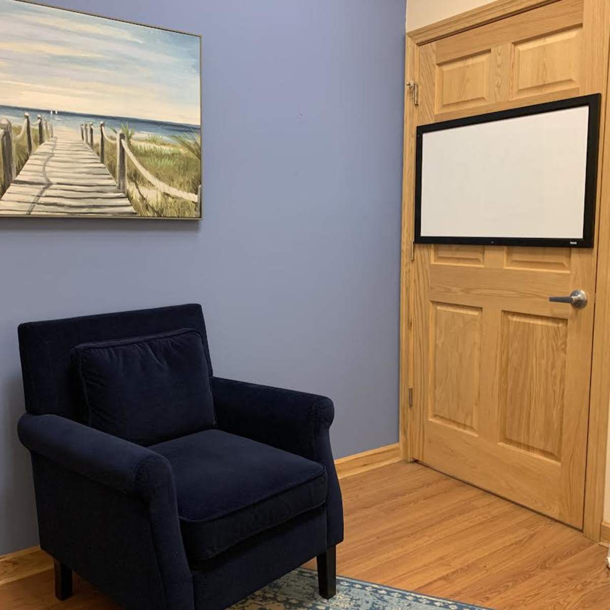 A room with a blue wall, a wooden door, a painting of a boardwalk leading to the beach, a blue armchair, and a blank wall-mounted screen.
