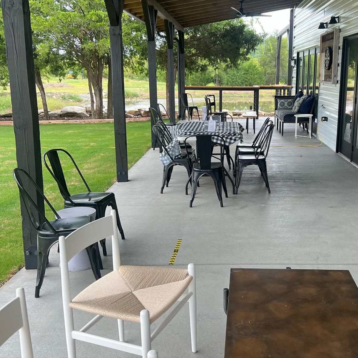 A shaded outdoor patio features a table set for dining, surrounded by black metal chairs, with additional seating options nearby.
