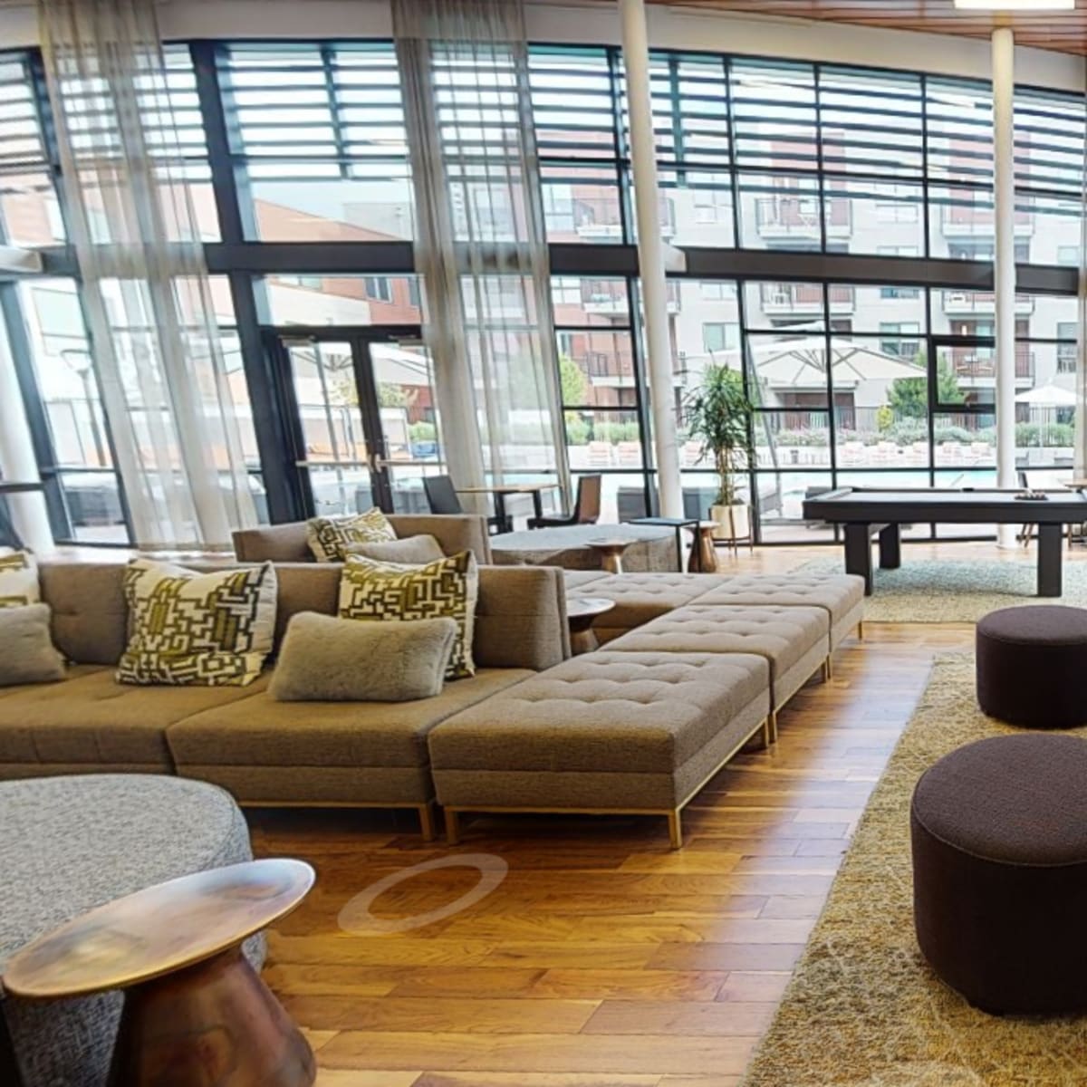 A modern lounge area with a sectional sofa, green chairs, round tables, and large windows overlooking an outdoor patio.