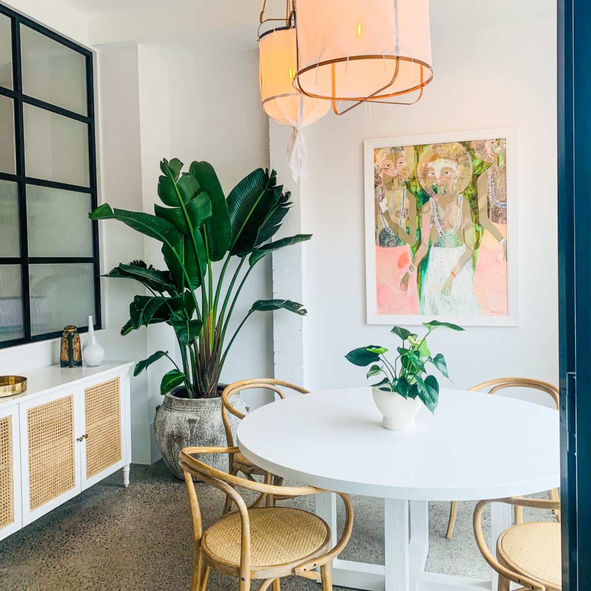 A dining area features a round white table surrounded by four rattan chairs, with large plants and artwork on the walls.