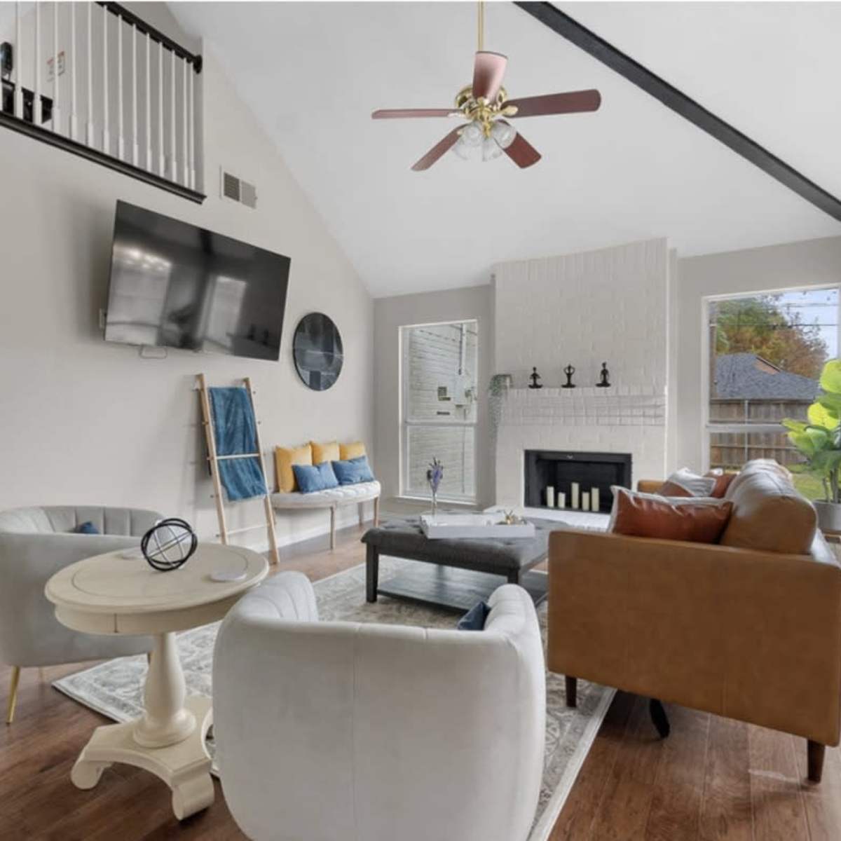 A bright living room features a large window, a flat-screen TV on the wall, and a mix of modern furniture including a coffee table and two sofas.