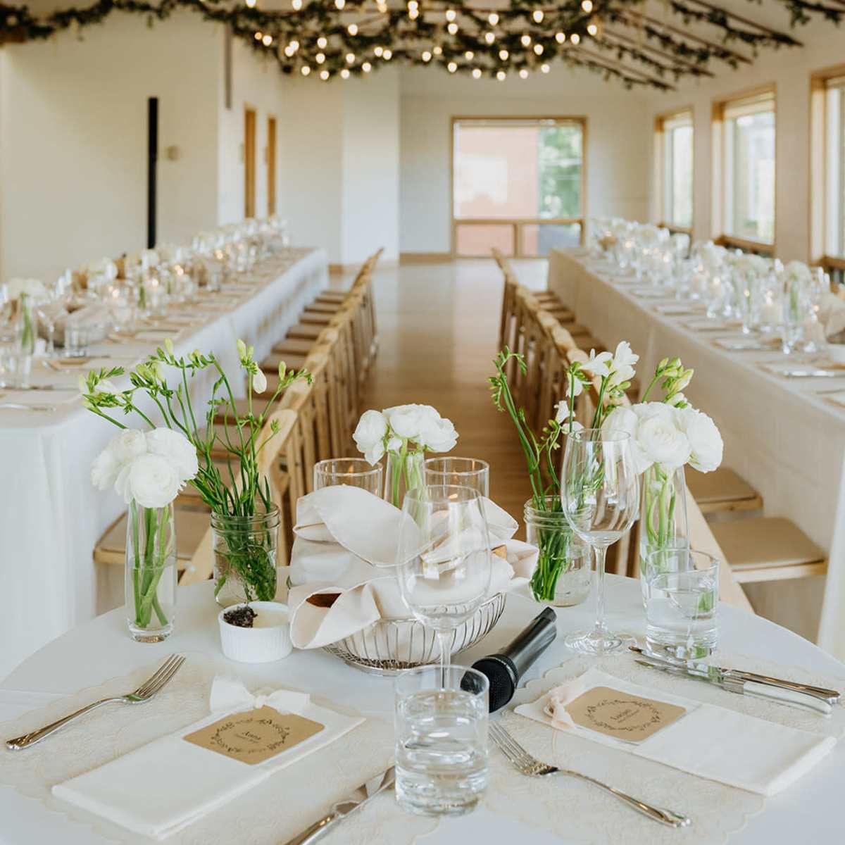 A beautifully arranged banquet hall features long tables with white linens, centerpieces of flowers and candles, surrounded by decorative greenery and soft lighting.