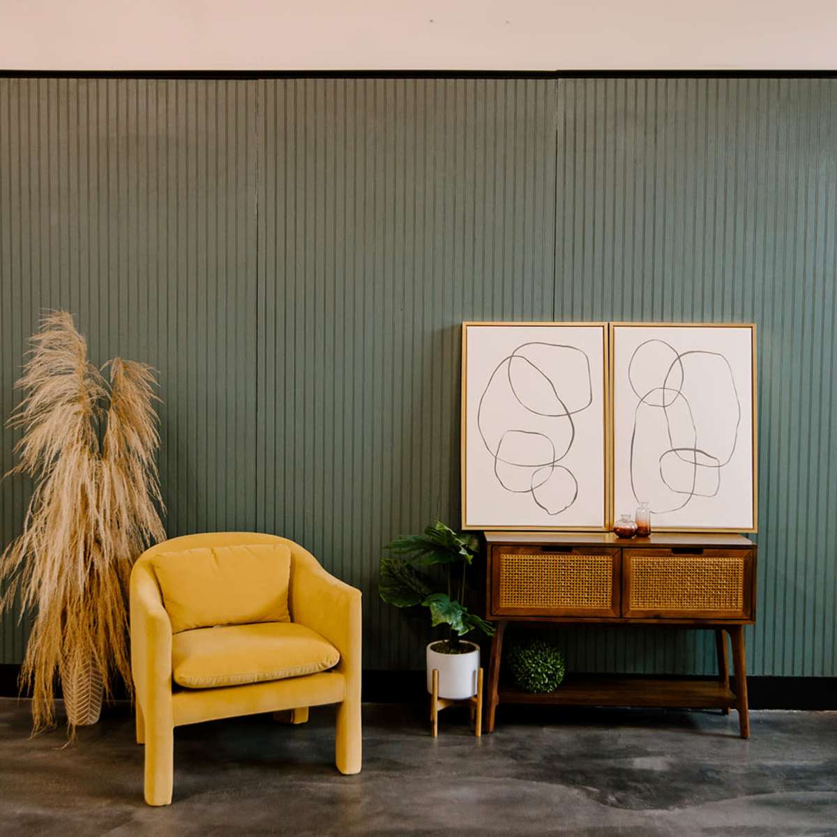 A yellow chair sits next to a woven console table with framed line art, accompanied by a tall decorative plant against a textured green wall.