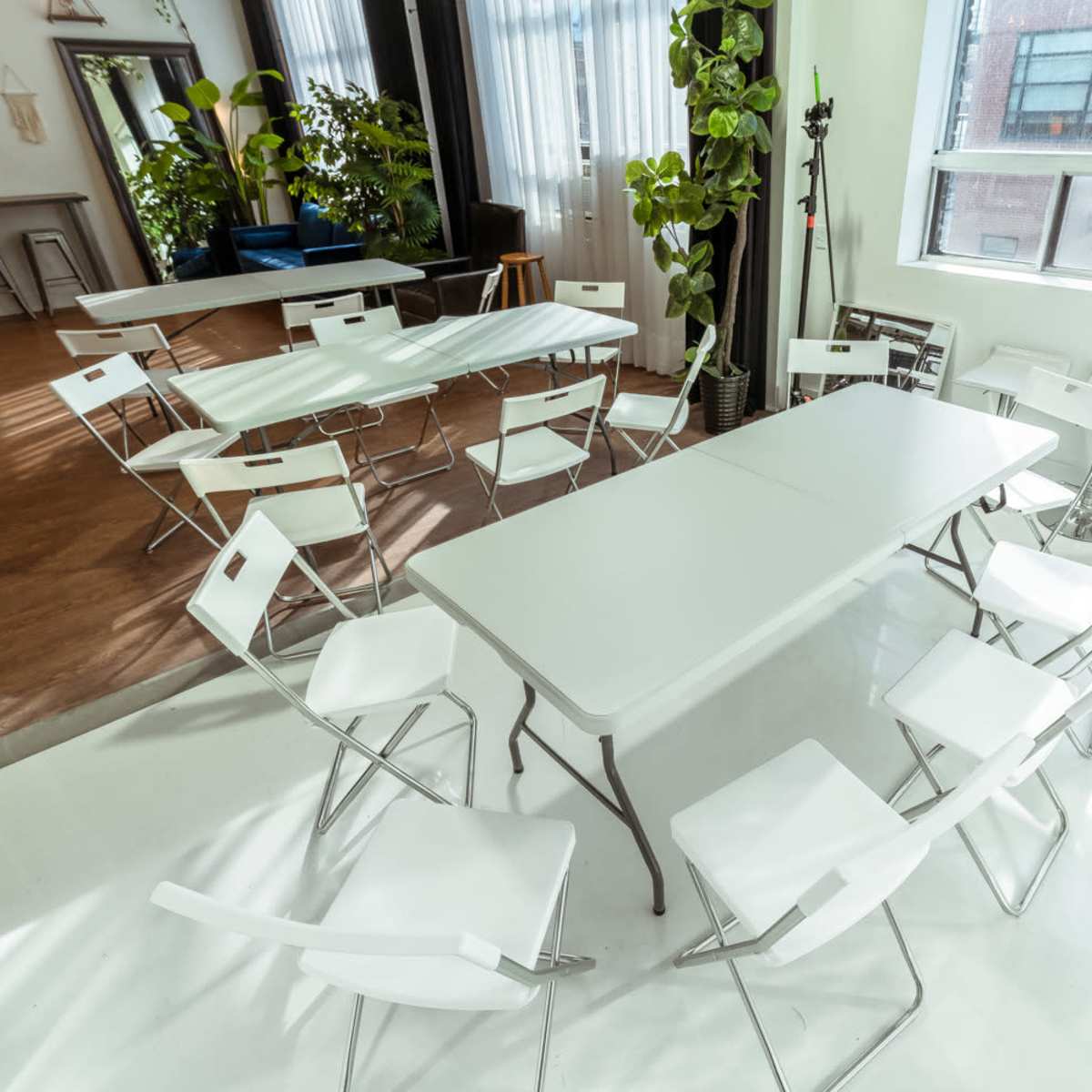 A bright room arranged with multiple folding tables and chairs, surrounded by large windows and greenery.