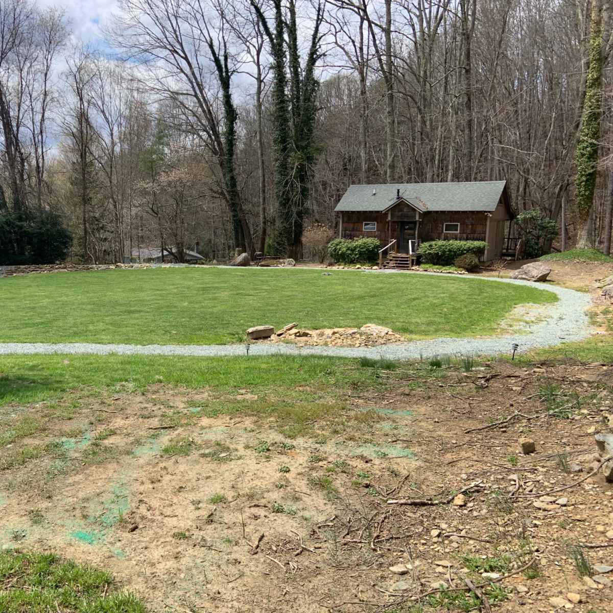 A grassy clearing with a gravel path leads to a wooden cabin surrounded by trees.