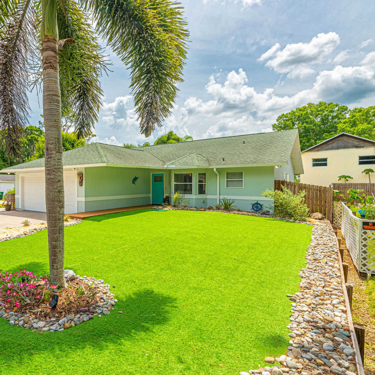 A single-story house with a light blue exterior is surrounded by well-maintained green lawn and decorative stones, featuring a palm tree in the foreground.