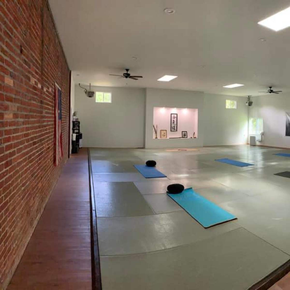 A spacious, well-lit studio with wooden floors, a large mat area, and yoga mats arranged on the floor.