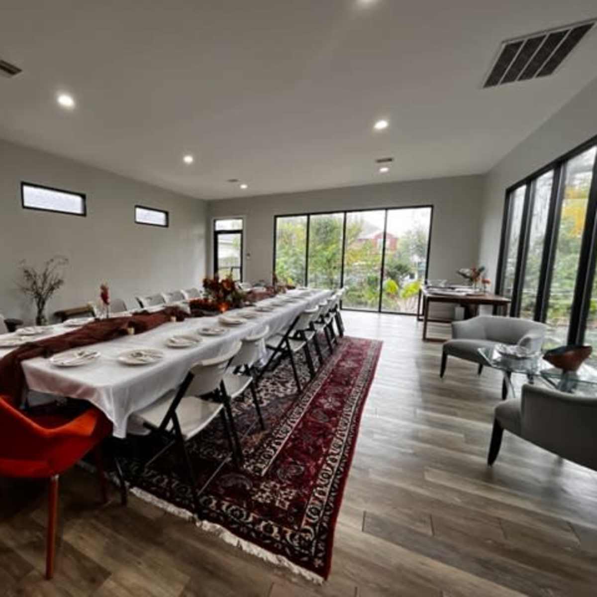 A long dining table with white linens is set for a meal in a spacious room featuring large windows and a view of greenery.