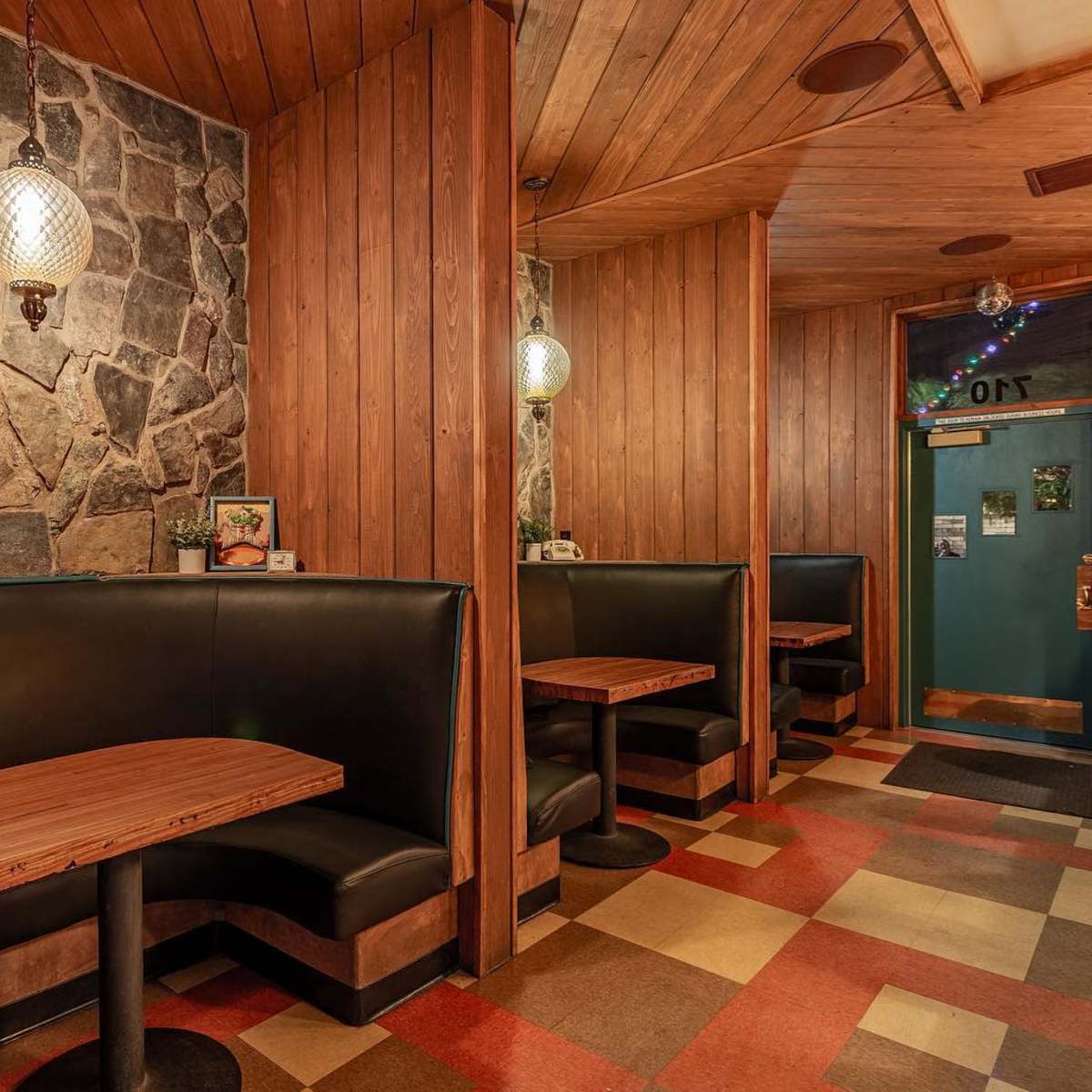 A cozy restaurant interior with wooden paneling, stone walls, and black booths featuring circular tables on a checkered floor.