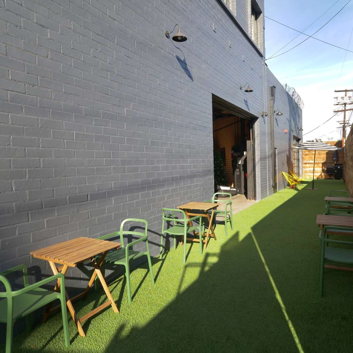 A narrow outdoor space with green artificial grass, wooden tables, and chairs along a brick wall and an open entrance to a building.