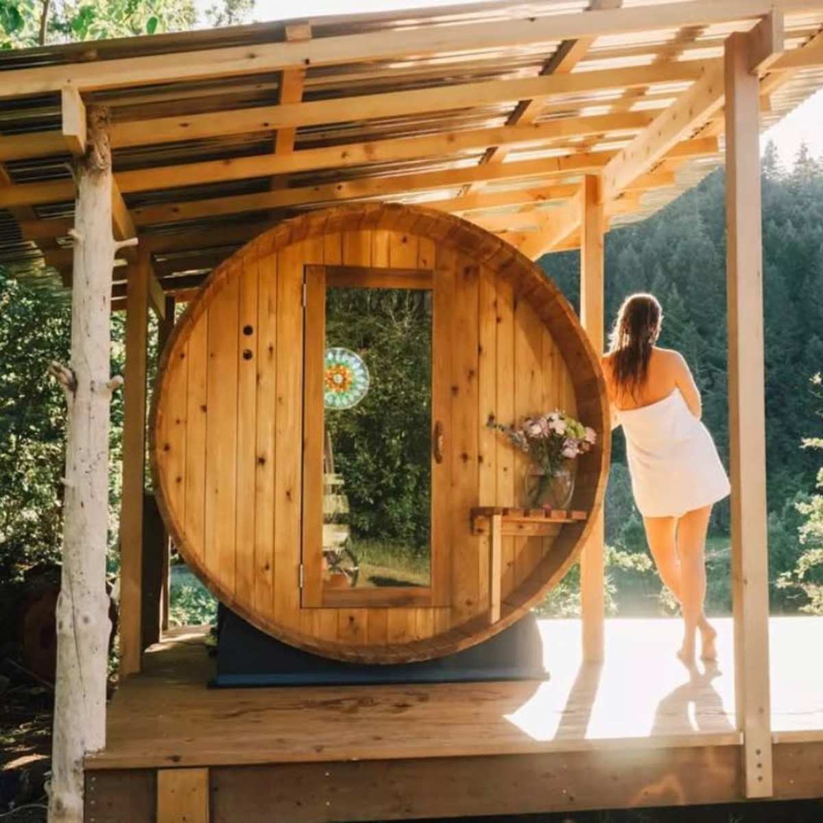 A woman stands on a deck next to a round wooden sauna, surrounded by trees and mountains.