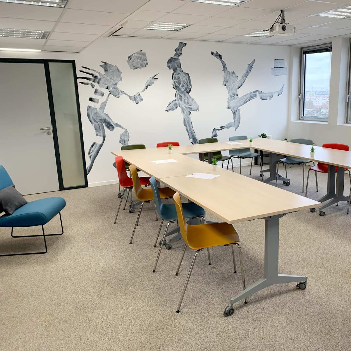 A spacious meeting room features multiple tables arranged in a U-shape, surrounded by colorful chairs and a mural of dancers on the wall.