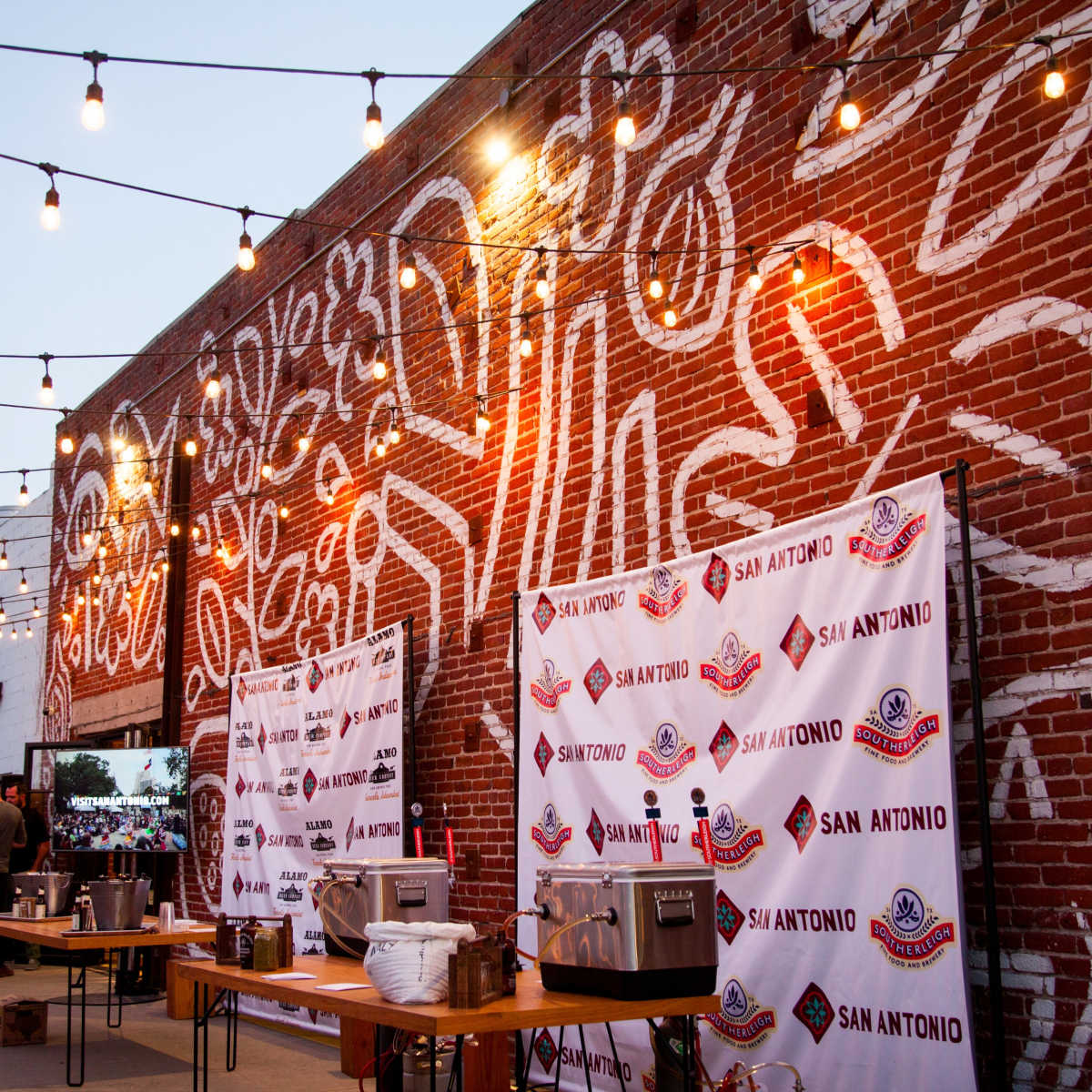 An outdoor event space with string lights above, featuring large murals on a brick wall and tables set up with catering equipment.