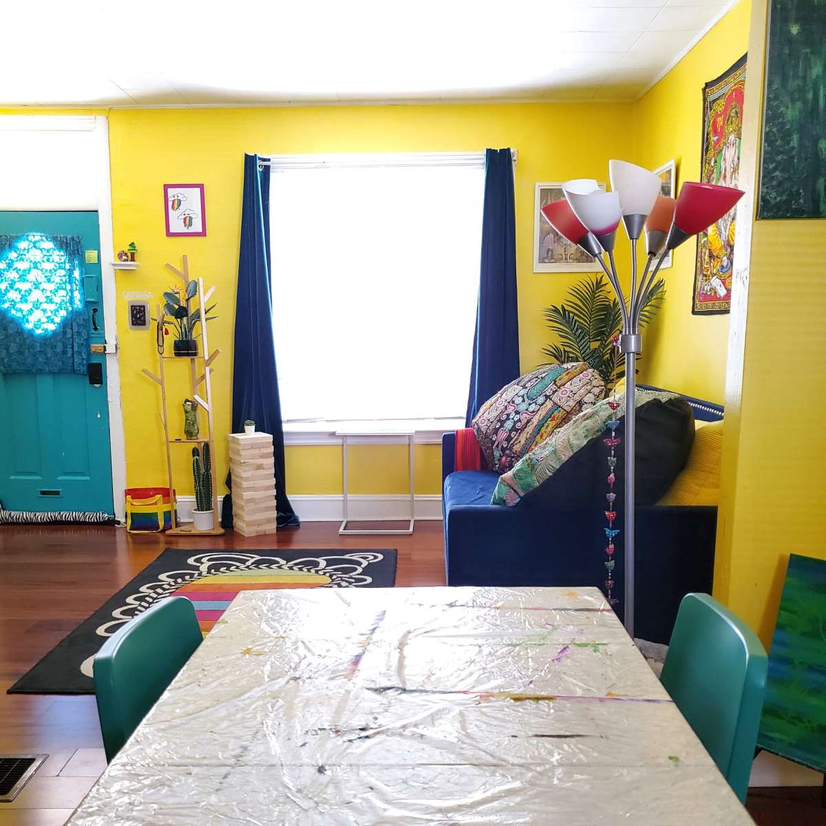 A bright yellow room with a table covered in plastic, a blue couch adorned with colorful pillows, and a window dressed with dark curtains.