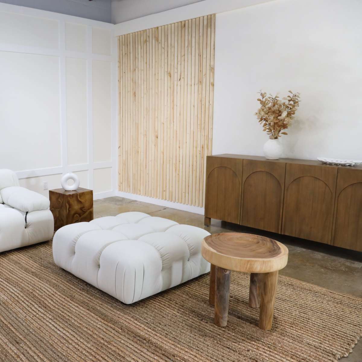A minimalist living room featuring a white sectional sofa, a tufted ottoman, a wooden side table, and a wooden cabinet against a textured wall.