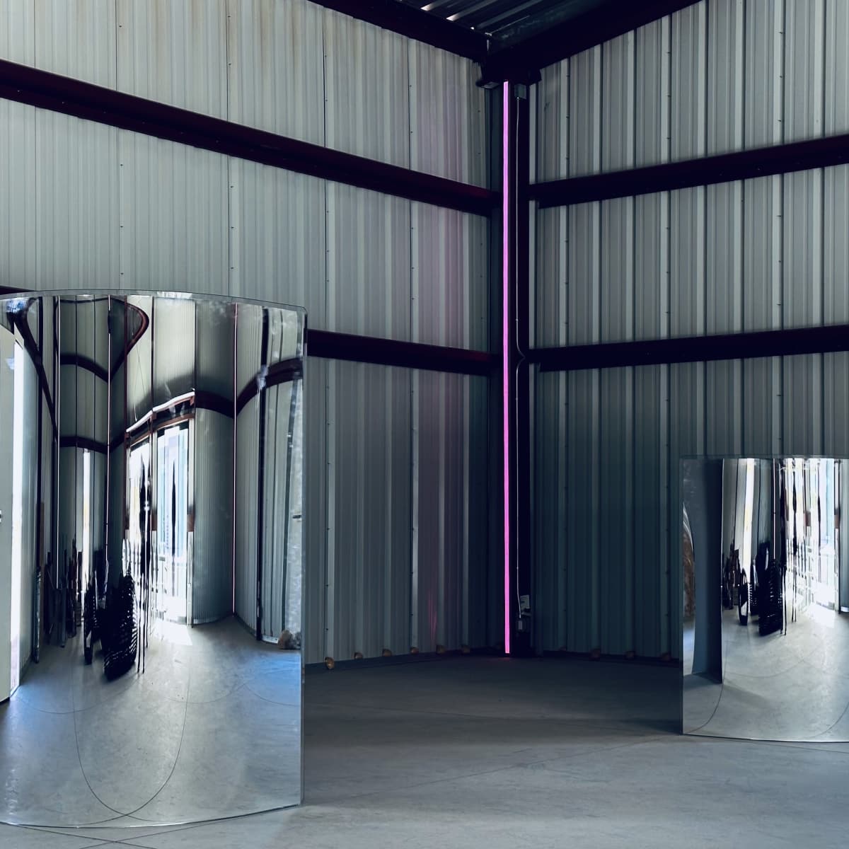 Two large, reflective metal sculptures stand in an industrial space with gray walls and a high ceiling.