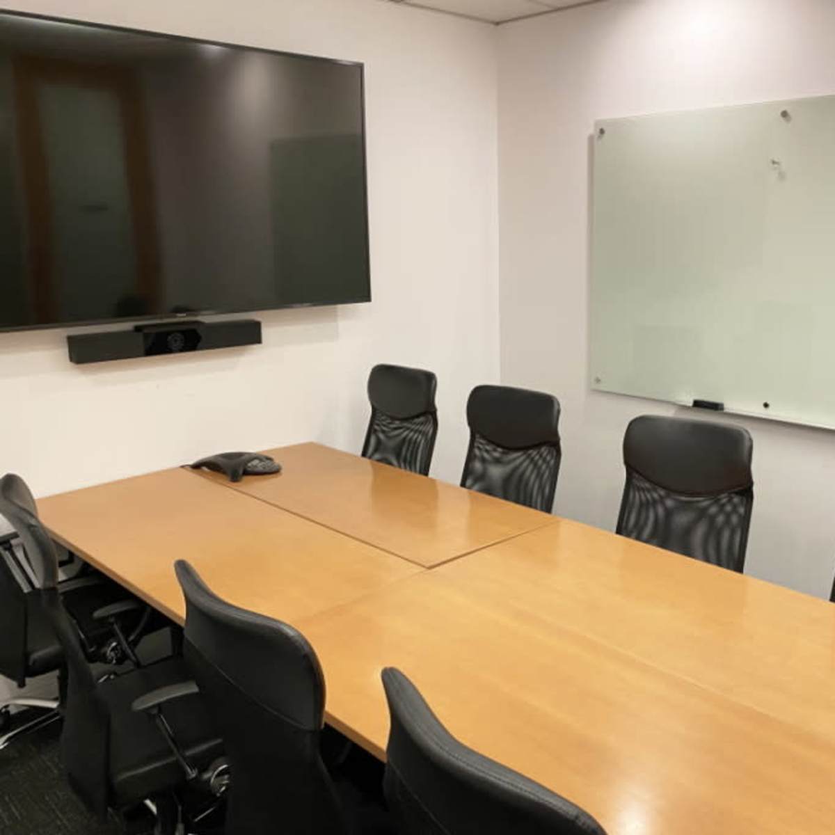 A conference room features a long wooden table bordered by black ergonomic chairs, a large screen mounted on the wall, and a whiteboard nearby.
