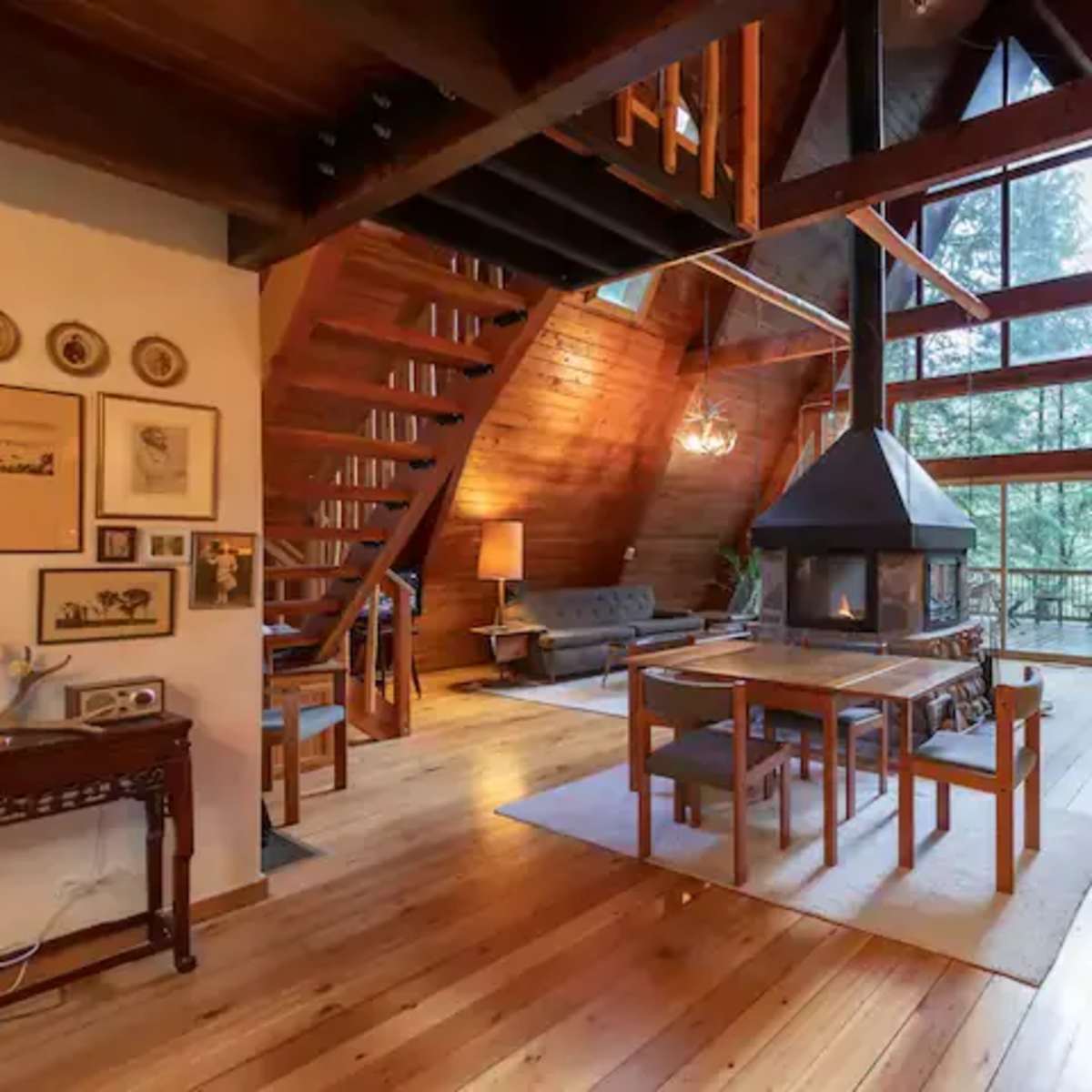 The interior of a wooden A-frame cabin features open spaces with a dining area, living space, and large windows overlooking trees.