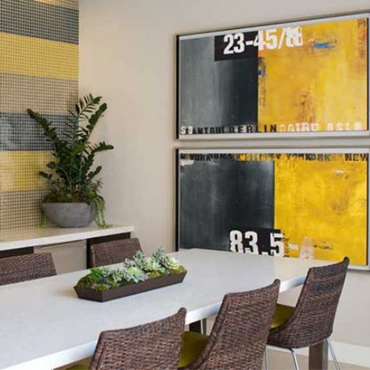 A modern dining area features a long table surrounded by woven chairs, with a mosaic wall and abstract art on display.