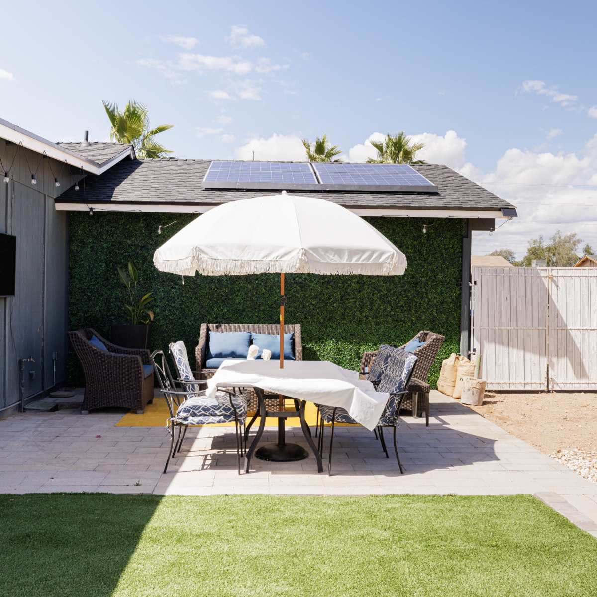 A patio area features a table with chairs under a large umbrella, surrounded by greenery and solar panels on the roof.