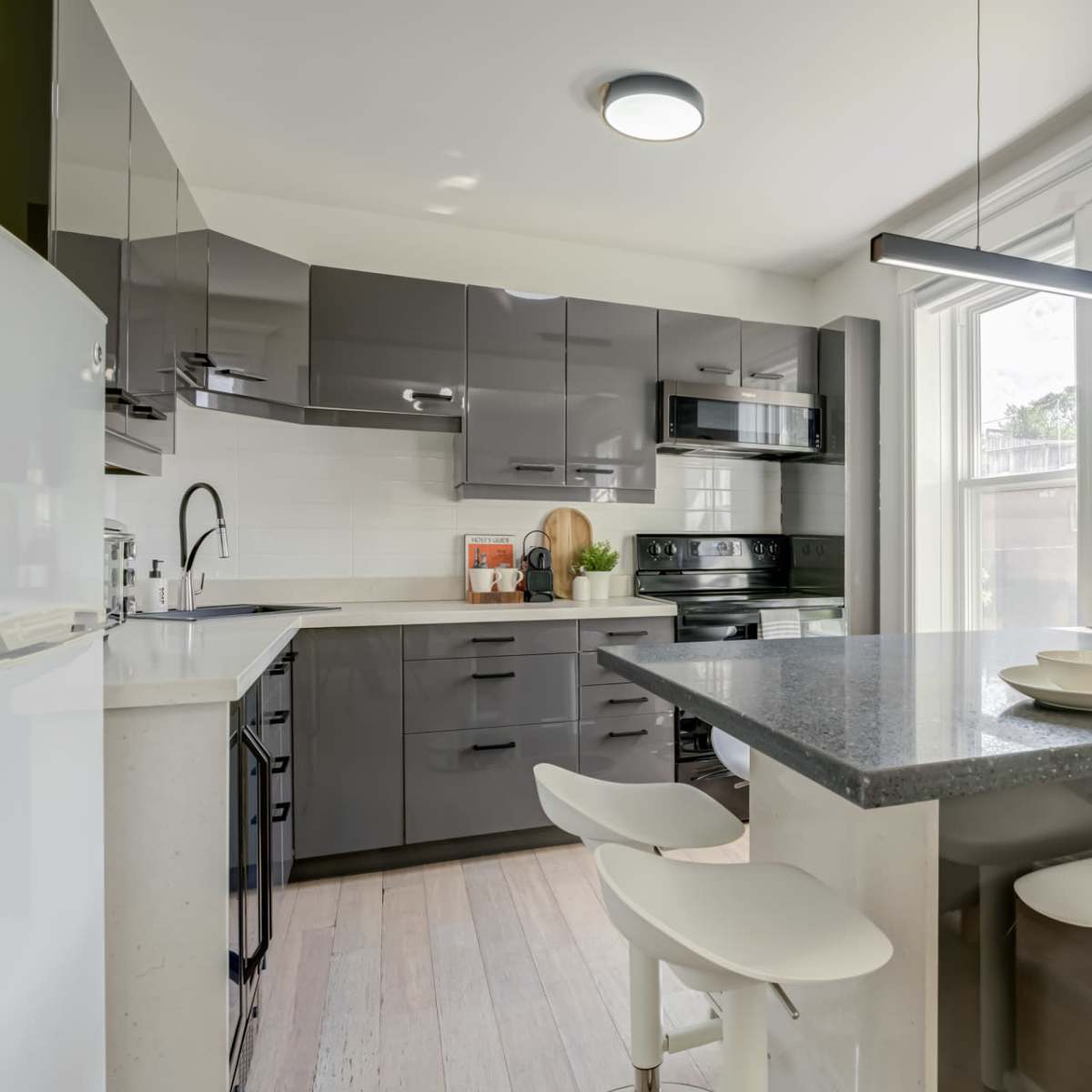 A modern kitchen with gray cabinets, a black countertop, and four white bar stools positioned around a small dining area.