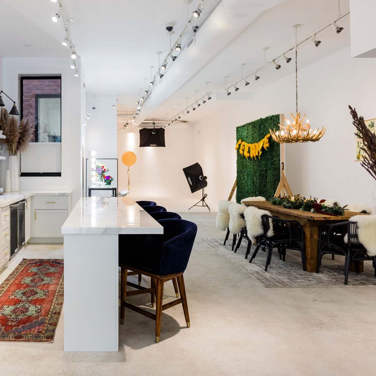 A modern, well-lit interior space featuring a kitchen area with a marble countertop, a dining area with a large wooden table, and decorative elements such as flowers and plants.