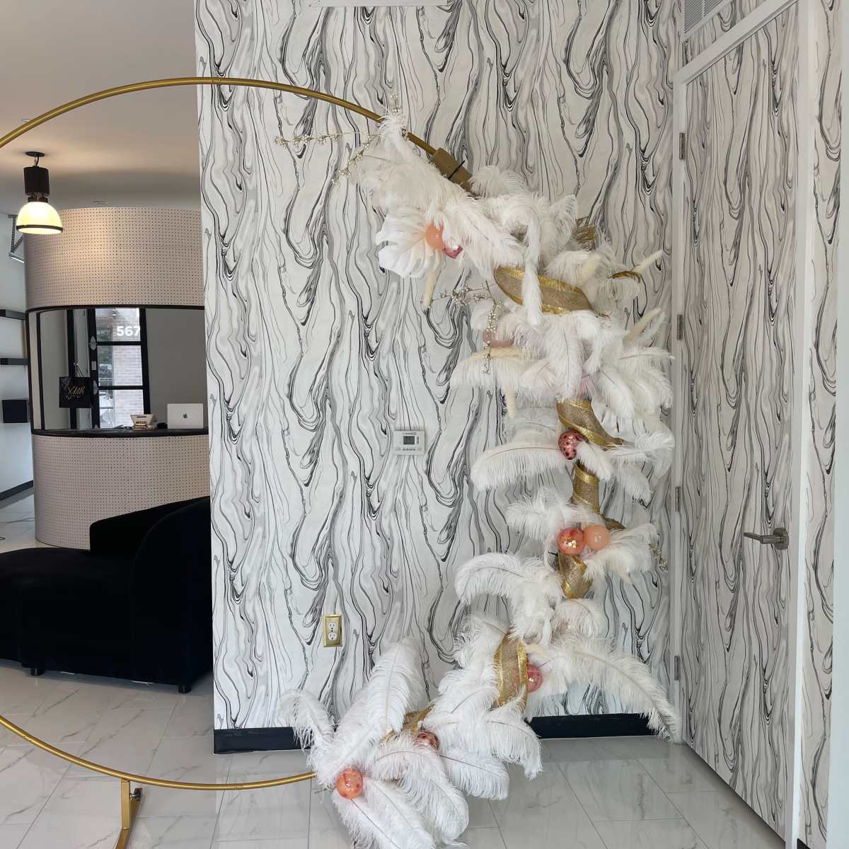 A tall decorative structure made of gold metal and white feathers is positioned near a reception area with a circular mirror and seating.