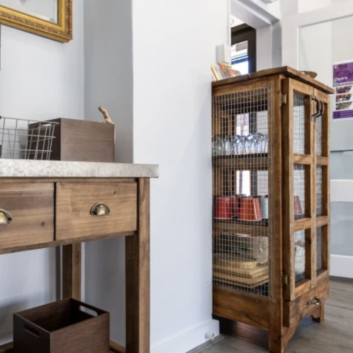 A wooden console table with drawers and a coffee maker is positioned against a wall, next to a wire-frame cabinet containing various items.