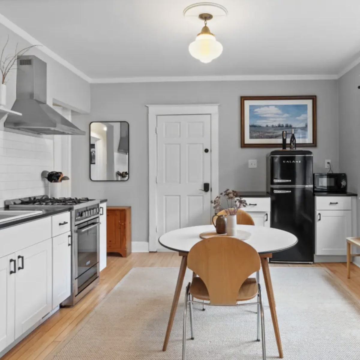A modern kitchen features a circular table with wooden chairs, a black refrigerator, and stainless steel appliances against light gray walls.