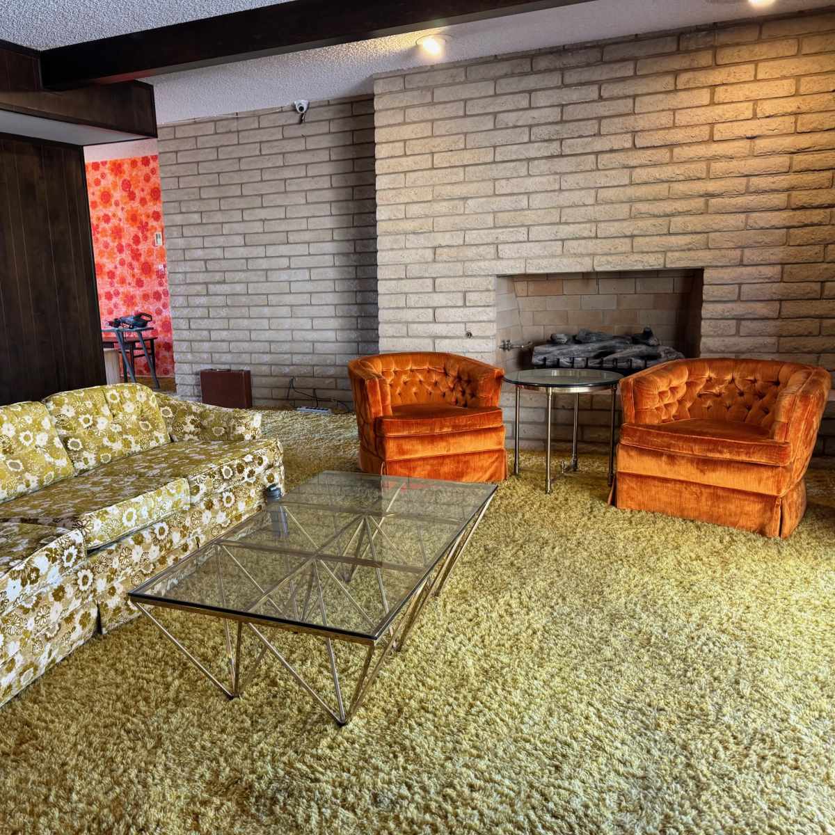 A living room with a floral-patterned sofa, two orange armchairs, a glass coffee table, and a stone fireplace against a textured wall.