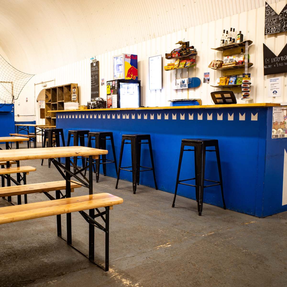 A spacious indoor bar area with wooden benches and a blue counter, featuring shelves of beverages and snacks in a well-lit environment.